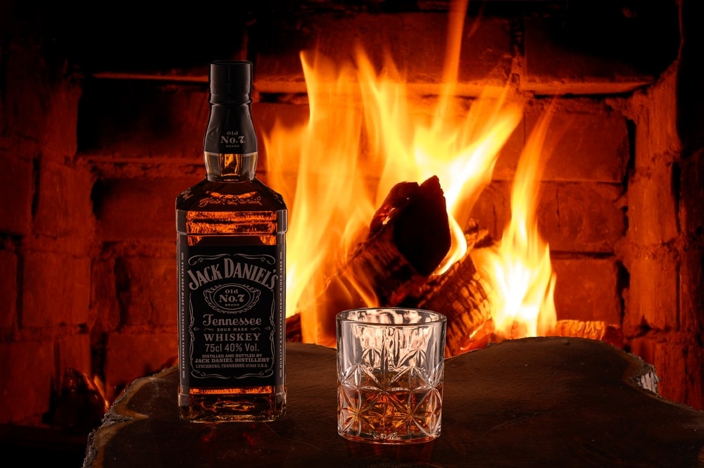 Whiskey Jack Daniel's Tennessee in a bottle and a glass of whiskey with ice in front of burning fireplace on a wooden natural cut of tree.